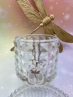 Collier en cristal de roche suspendu sur verre taillé, accompagné de libellules dorées – symbole de lumière, pureté et énergie universelle